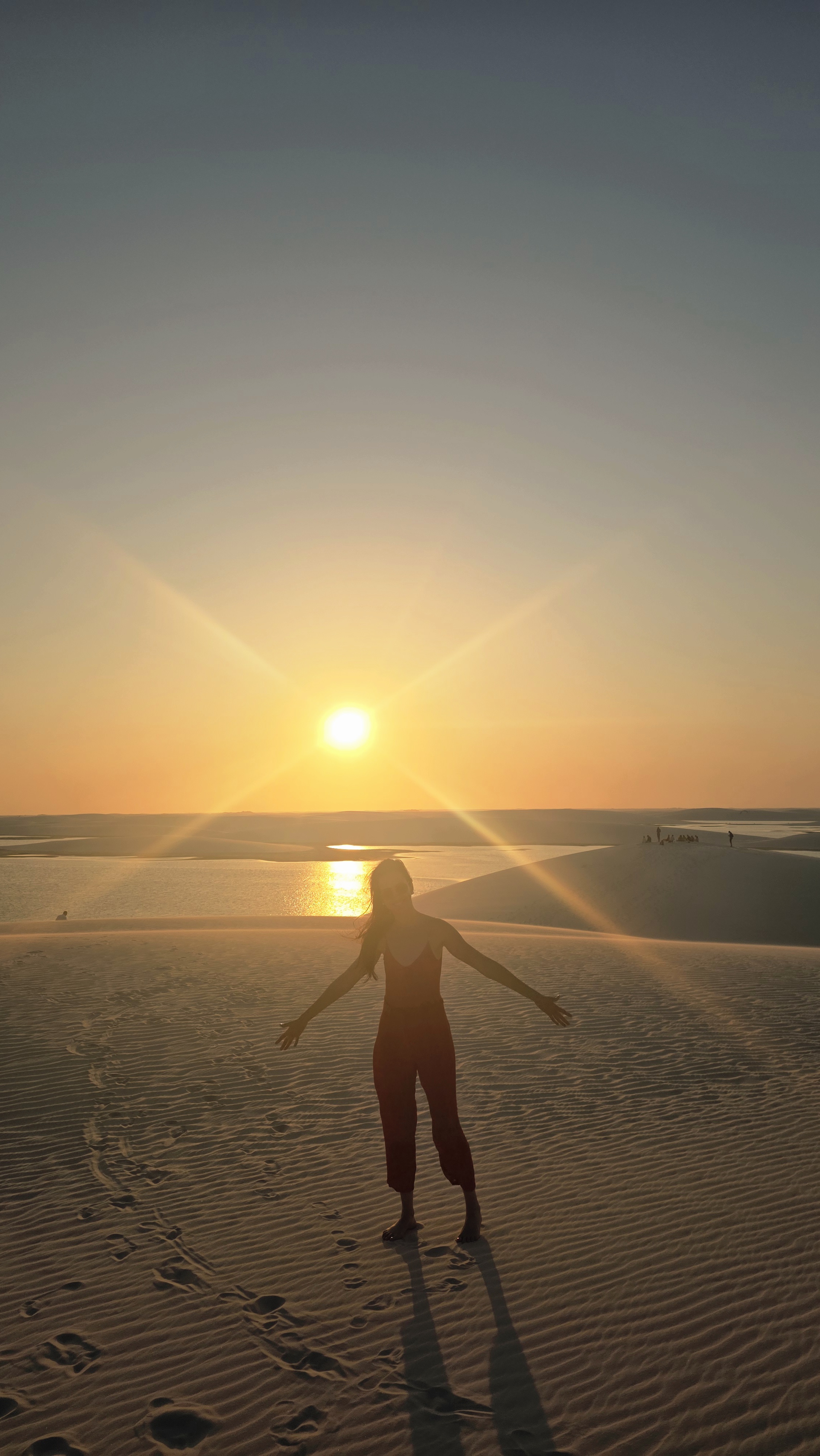 Sabrina em contraluz, de braços abertos no alto de uma duna, com o sol baixo atrás